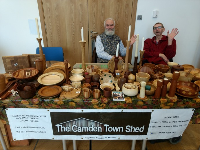 Photo of two Shed members, sat at a table of wooden bowls, and cups, waving